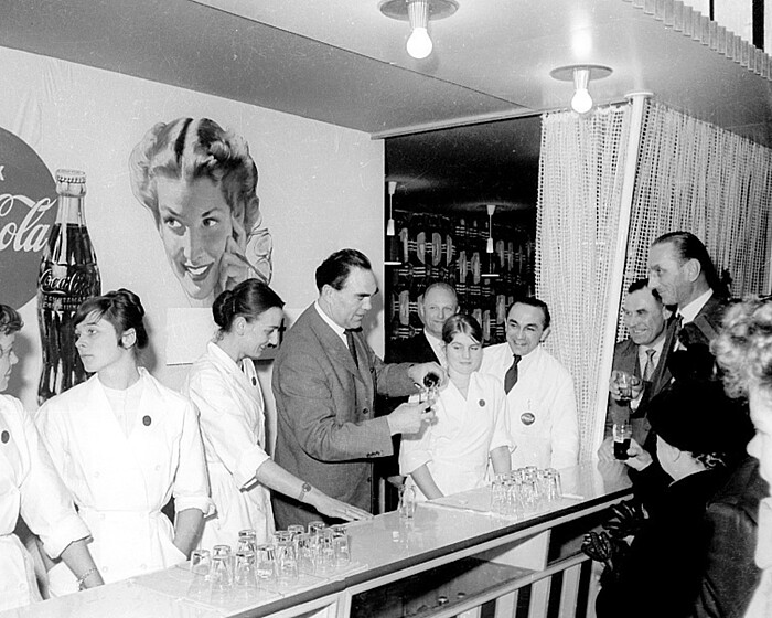A Coca-Cola exhibition booth from 1958, featuring a counter, glasses, and visitors during a product demonstration.