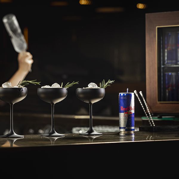 Bartresen mit drei Cocktailgläsern und einer Red Bull Dose, im Hintergrund weitere Red Bull-Dosen und eine Barkeeperin