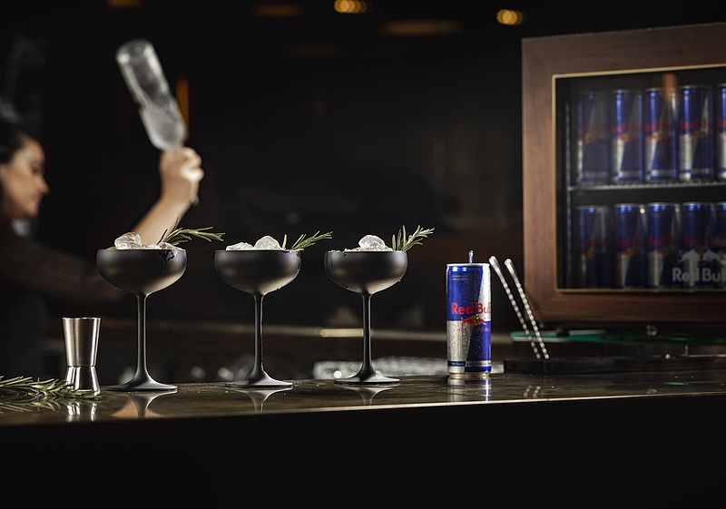 Bartresen mit drei Cocktailgläsern und einer Red Bull Dose, im Hintergrund weitere Red Bull-Dosen und eine Barkeeperin