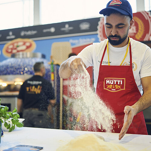 Mann mit blauer Kappe und roter Schürze arbeitet mit Mehl und Pizzateig.