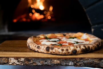 Eine Mozzarellapizza liegt auf einer Holzplatte vor einem Ofen.