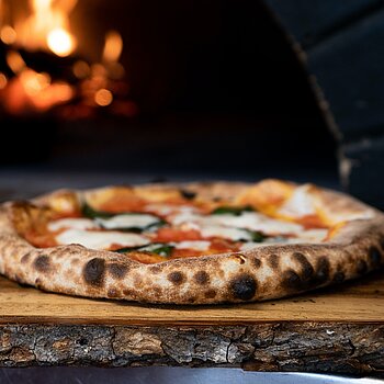 Eine Mozzarellapizza liegt auf einer Holzplatte vor einem Ofen.