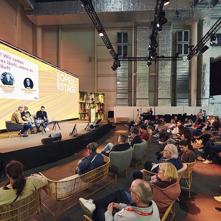 Eine große offene Veranstaltungsfläche mit einer Bühne, auf der zwei Personen in Sesseln sitzen und ein Gespräch vor Publikum führen. Auf einer großen Leinwand hinter ihnen ist der Titel einer Talkrunde zu sehen. Vor der Bühne sitzen zahlreiche Besucherinnen und Besucher in Sesseln und auf Stühlen und hören der Diskussion zu. Die Umgebung wirkt modern, mit Bühnenlicht, Pflanzenwänden und hoher Hallendecke.