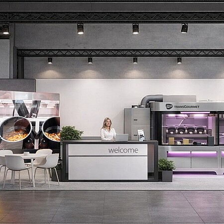 Messestand in modernem, offenen Design. In der Mitte steht ein Empfangstresen mit der Aufschrift „welcome“, hinter dem eine Person steht. Links befinden sich mehrere kleine Tische mit weißen Stühlen sowie eine große Wand mit einem Küchenmotiv. Rechts sind professionelle Gastronomiegeräte und Automaten mit lila Beleuchtung zu sehen, darüber das Logo „food@service“. Daneben stehen zwei blaue Informations- oder Bestellterminals.