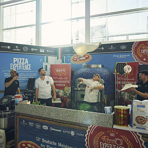 Fünf Männer sind zu sehen, zwei davon formen mittels Pizza Akrobatik den Teig.