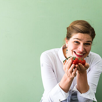 Antje de Vries steht vor einem hellgrünen Hintergrund und hält lachend kleine Tomaten in die Kamera 