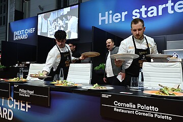Mehrere Köche in weißer Arbeitskleidung servieren Teller mit angerichteten Speisen an einem Jurytisch beim „Next Chef Award“. Vor den weißen Stühlen stehen Namensschilder, unter anderem für Philipp Vogel („Orania Berlin“) und Cornelia Poletto („Restaurant Cornelia Poletto“). Im Hintergrund sind ein großer Bildschirm mit einer Live-Küchenaufnahme und die Worte „Hier starte…“ auf einer blauen Wand zu sehen.