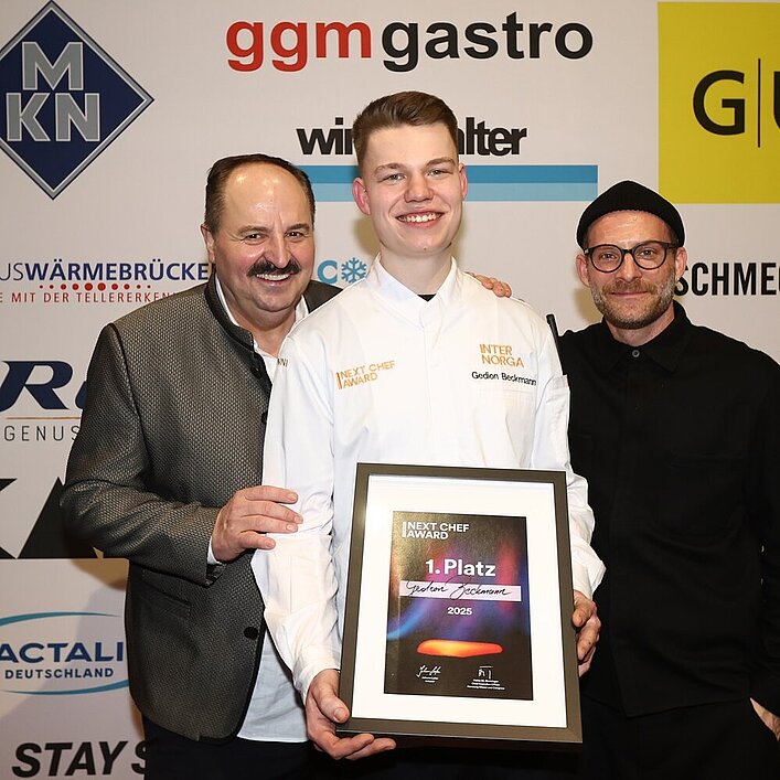 Der Gewinner des Next Chef Awards 2025, Gedion Beckmann, hält stolz seine Siegerurkunde, während Sternekoch Johann Lafer (links) und Moderator Sebastian E. Merget (rechts) ihn gratulierend flankieren.