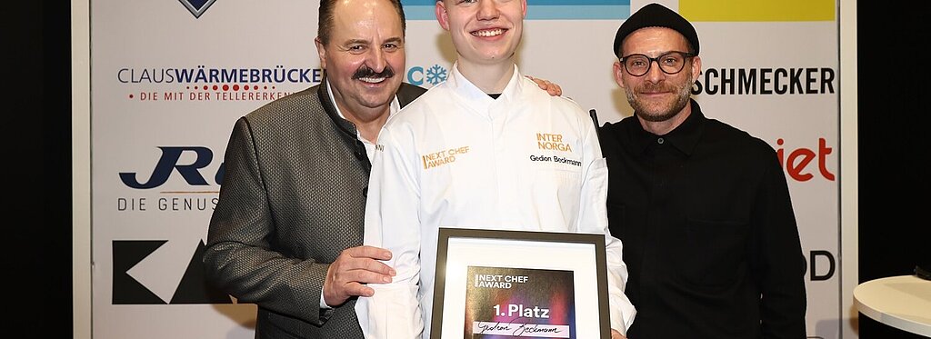 Der Gewinner des Next Chef Awards 2025, Gedion Beckmann, hält stolz seine Siegerurkunde, während Sternekoch Johann Lafer (links) und Moderator Sebastian E. Merget (rechts) ihn gratulierend flankieren.
