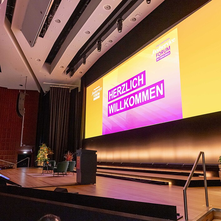 Großer Konferenzsaal mit Bühne und riesiger Leinwand. Auf dem Bildschirm steht „HERZLICH WILLKOMMEN“ vor gelb-pinkem Hintergrund des International Foodservice Forum. Die Bühne ist mit Sesseln, Rednerpult und tropischen Pflanzen dekoriert, während links Zuschauer im Auditorium sitzen.