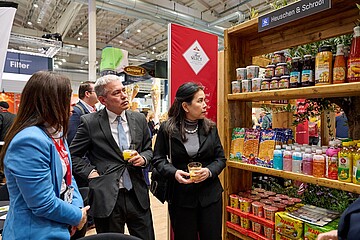 Mehrere Personen stehen auf einer Messe vor einem Ausstellungsregal mit Lebensmitteln und Getränken des Unternehmens Heuschen & Schroo und unterhalten sich, während sie Getränke in der Hand halten.