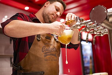 Eine Person in kariertem Hemd und Leder-Schürze zapft Bier an einem Tresen und füllt ein Glas, während Schaum über den Rand läuft. Mehrere Zapfhähne sind im Hintergrund zu sehen.