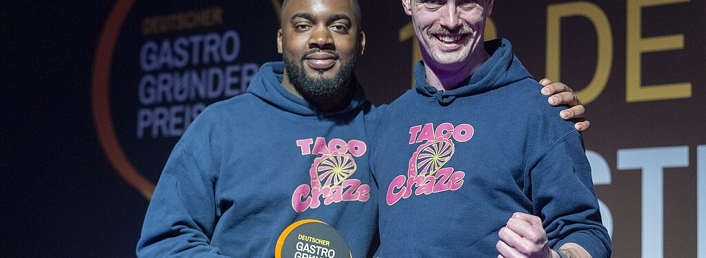Zwei Personen in blauen „TACO Craze“-Hoodies stehen nebeneinander und halten eine runde Auszeichnung mit der Aufschrift „DEUTSCHER GASTRO GRÜNDER PREIS“.