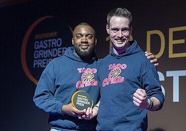 Zwei Personen in blauen „TACO Craze“-Hoodies stehen nebeneinander und halten eine runde Auszeichnung mit der Aufschrift „DEUTSCHER GASTRO GRÜNDER PREIS“.