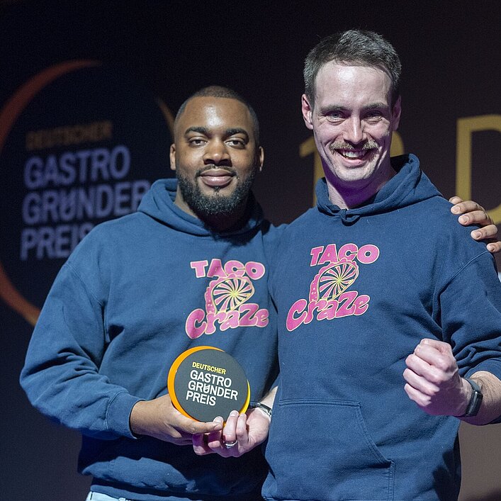 Zwei Personen in blauen „TACO Craze“-Hoodies stehen nebeneinander und halten eine runde Auszeichnung mit der Aufschrift „DEUTSCHER GASTRO GRÜNDER PREIS“.