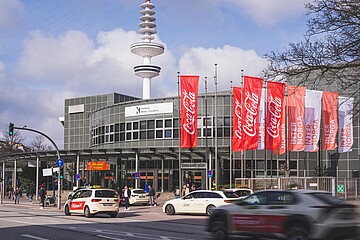 Eine Straßenansicht vor einem Messeeingang mit mehreren vorbeifahrenden Autos. Am Gebäude hängen rote und weiße Fahnen mit Marken- und Messebeschriftung, im Hintergrund steht ein hoher Turm.
