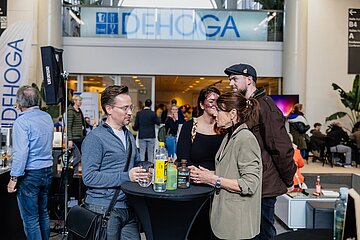 Eine Gruppe von Menschen steht an einem Stehtisch bei einer DEHOGA-Veranstaltung und unterhält sich. Im Hintergrund sind das DEHOGA-Logo und weitere Messebesucher zu sehen. Auf dem Tisch stehen verschiedene Getränke.