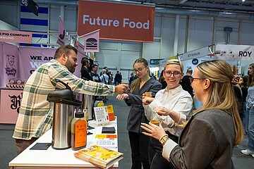 An einem Messebereich mit dem Schild „Future Food“ probieren Besucherinnen und Besucher Produkte an einem Stand. Auf dem Tisch stehen Getränke und Informationsmaterial, während eine Person hinter dem Stand Probierportionen ausgibt.
