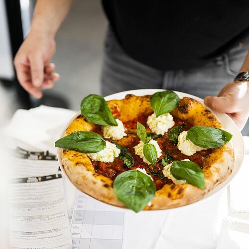 Ein Mann hält eine Pizza in der Hand, welche mit Mozzarella und Basilikum belegt ist. 