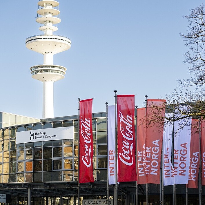Außenansicht des Hamburg Messe und Congress Gebäudes bei klarem Himmel. Vor dem Eingang stehen mehrere rote und weiße Fahnen mit Coca-Cola- und INTERNORGA‑Branding. Im Hintergrund ragt der markante Heinrich-Hertz-Turm in den Himmel.