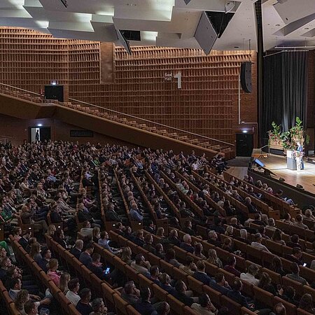 „Eine voll besetzte Konferenzhalle mit moderner Holzarchitektur. Auf der Bühne spricht eine Person am Rednerpult, während ihre Präsentation auf einer großen Leinwand im Hintergrund projiziert wird.“
