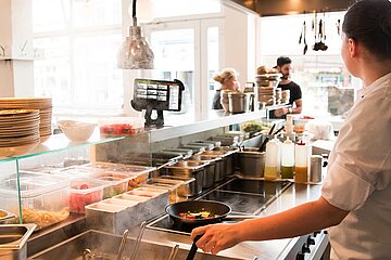 Blick in die Küche eines Restaurants, ein Koch steht mit einer Pfanne am Herd