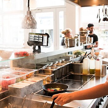 Blick in die Küche eines Restaurants, ein Koch steht mit einer Pfanne am Herd