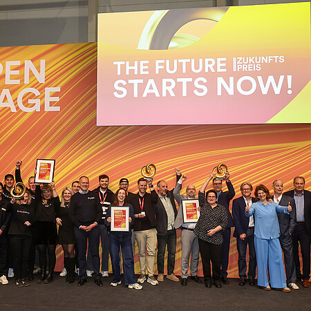 Gruppenfoto einer größeren Gruppe von ausgezeichneten Personen auf einer Bühne der Internorga. Sie halten Urkunden und Trophäen vor einer orange-gelben Bühnenkulisse mit der Aufschrift ‚Open Stage‘ und ‚The Future Starts Now!