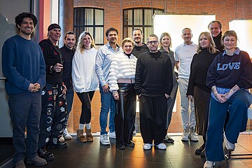 Eine Gruppe von Menschen steht für ein gemeinsames Foto in einem Innenraum vor einer Backsteinwand. Einige Personen stehen, andere sitzen, und die Gruppe trägt überwiegend legere Kleidung. Helle Studiobeleuchtung befindet sich hinter ihnen und sorgt für eine freundliche, ausgeleuchtete Szene.