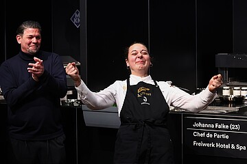 Eine Person in Kochkleidung und dunkler Schürze steht auf einer Bühne und hebt beide Arme leicht an. Daneben steht eine weitere Person und klatscht. Im Hintergrund sind Küchenelemente zu sehen.