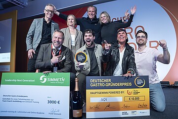 Eine Gruppe von neun Personen posiert auf einer Bühne vor einem großen orange‑weißen Hintergrund mit der Aufschrift „Deutscher Gastro‑Gründerpreis“. Vor der Gruppe stehen zwei große symbolische Schecks: links ein grüner Scheck über 3.000 Euro für einen Ausbildungsplatz bei Simmet‑Training, rechts ein orangefarbener Hauptgewinn‑Scheck über 10.000 Euro mit dem Gewinnernamen „MOI“. Davor steht zudem eine große Flasche Sekt. Eine Person in der Mitte hält eine runde Auszeichnungstrophäe hoch. Die Gruppe zeigt teils jubelnde Gesten und wirkt wie bei einer Siegerehrung. Geben Sie Ihr Feedback zu BizChat ab.