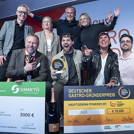 Eine Gruppe von neun Personen posiert auf einer Bühne vor einem großen orange‑weißen Hintergrund mit der Aufschrift „Deutscher Gastro‑Gründerpreis“. Vor der Gruppe stehen zwei große symbolische Schecks: links ein grüner Scheck über 3.000 Euro für einen Ausbildungsplatz bei Simmet‑Training, rechts ein orangefarbener Hauptgewinn‑Scheck über 10.000 Euro mit dem Gewinnernamen „MOI“. Davor steht zudem eine große Flasche Sekt. Eine Person in der Mitte hält eine runde Auszeichnungstrophäe hoch. Die Gruppe zeigt teils jubelnde Gesten und wirkt wie bei einer Siegerehrung. Geben Sie Ihr Feedback zu BizChat ab.